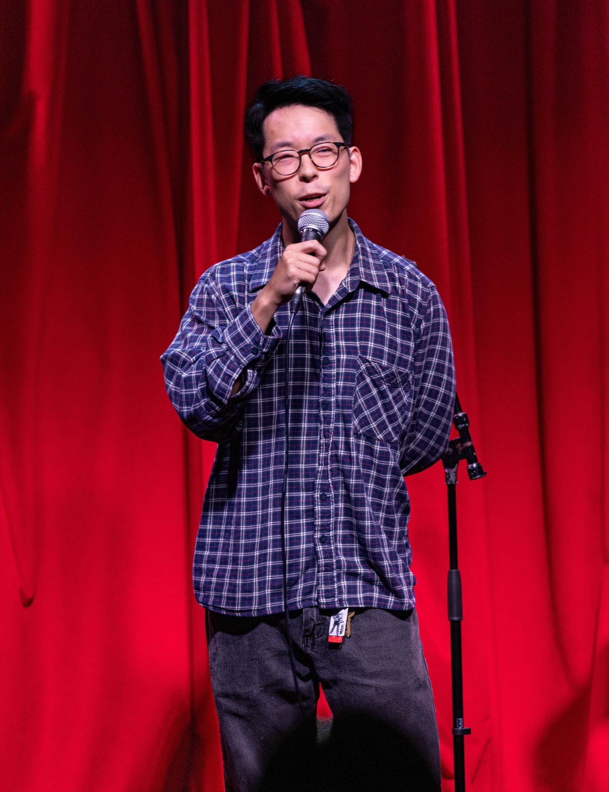A performer with a microphone in front of the red curtain at Baklava Comedy.